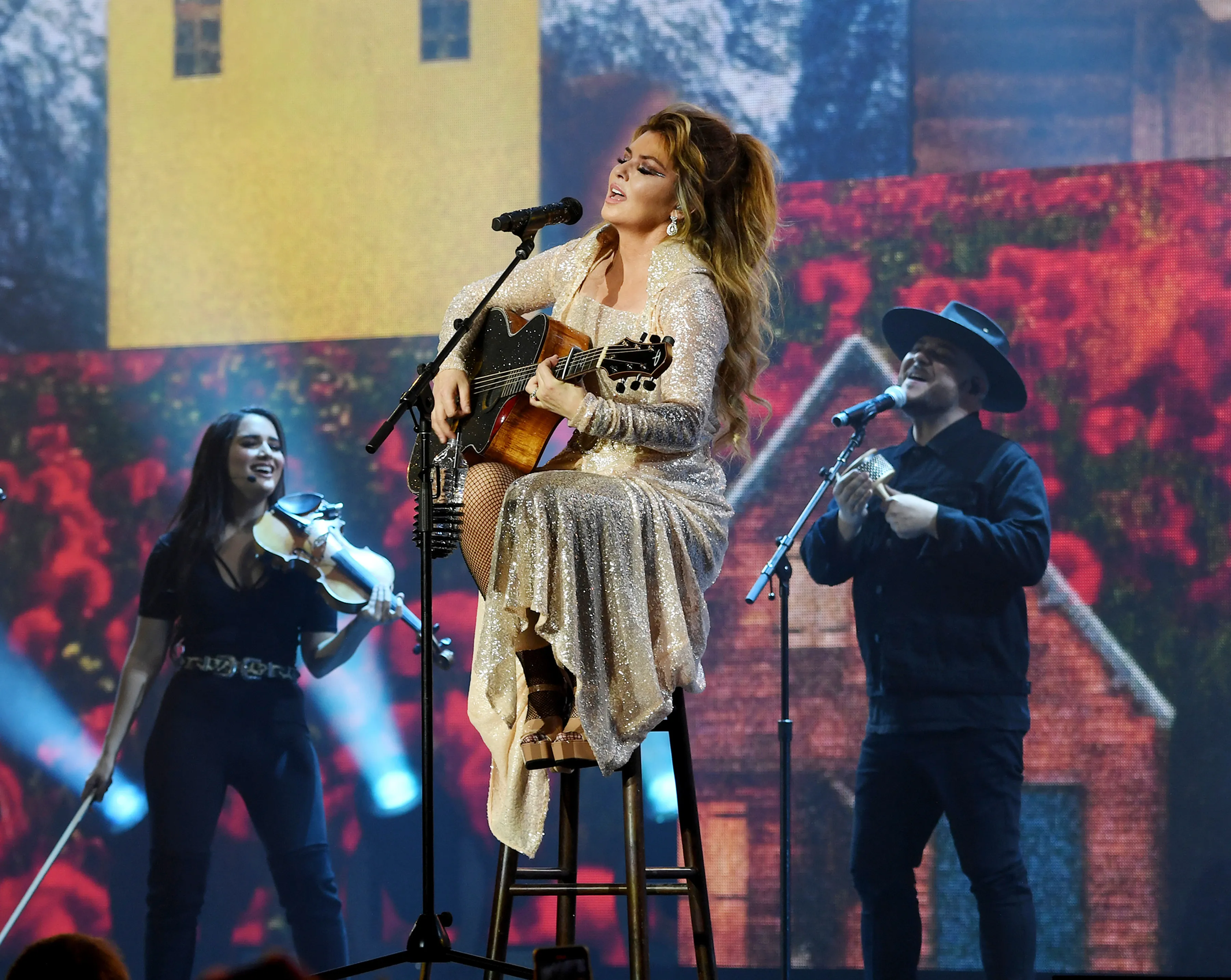 Shania Twain performs while seated on a stool, playing an acoustic guitar and singing into a microphone, flanked by a female violinist and a male vocalist.