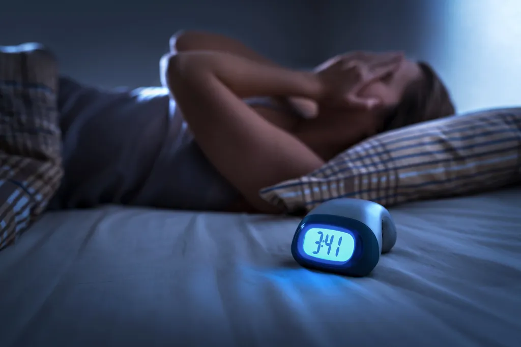 A woman lying awake in bed with her hands covering her face, next to an alarm clock displaying 3:41.