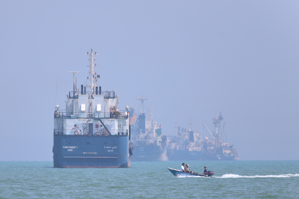 A small motorboat with three people passing large anchored vessels in the Strait of Hormuz.