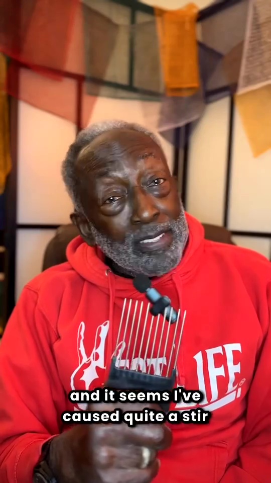 Garrett Morris in a red hoodie holding an afro pick with a microphone attached, addressing a viral moment.