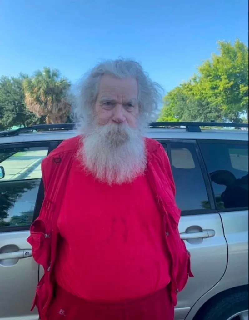 Man named Santa Claus, arrested for alleged child exploitation, stands in a red shirt and jacket with a full white beard and long white hair.