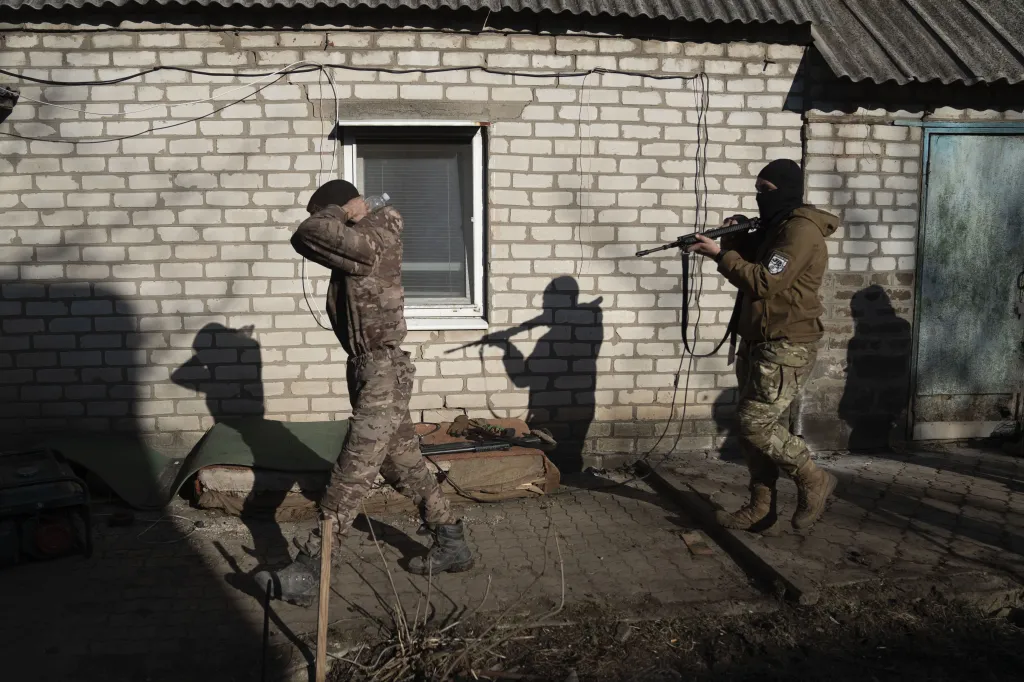 A soldier in camouflage, holding a water bottle, walks past a soldier in a black balaclava holding a rifle, next to a brick building.