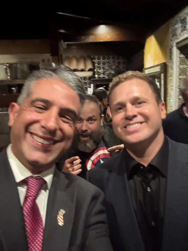 Two men smiling at the camera, with a third bearded man in the background smiling and pointing.