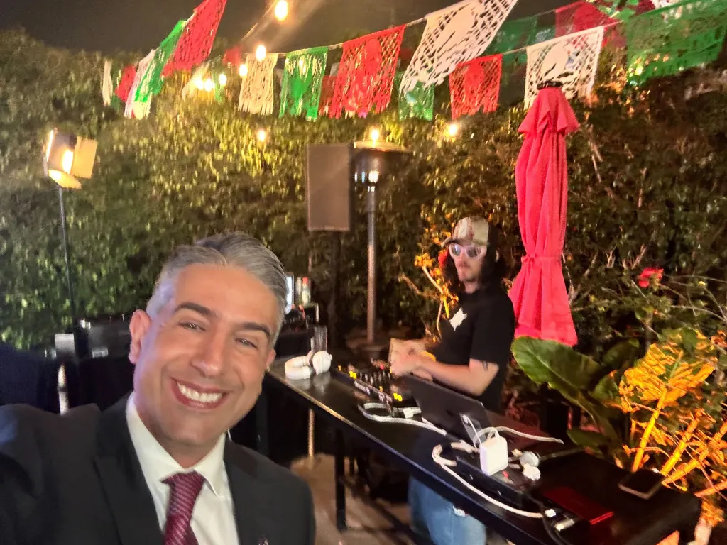 Spencer Pratt smiling with a DJ at a party with papel picado banners.