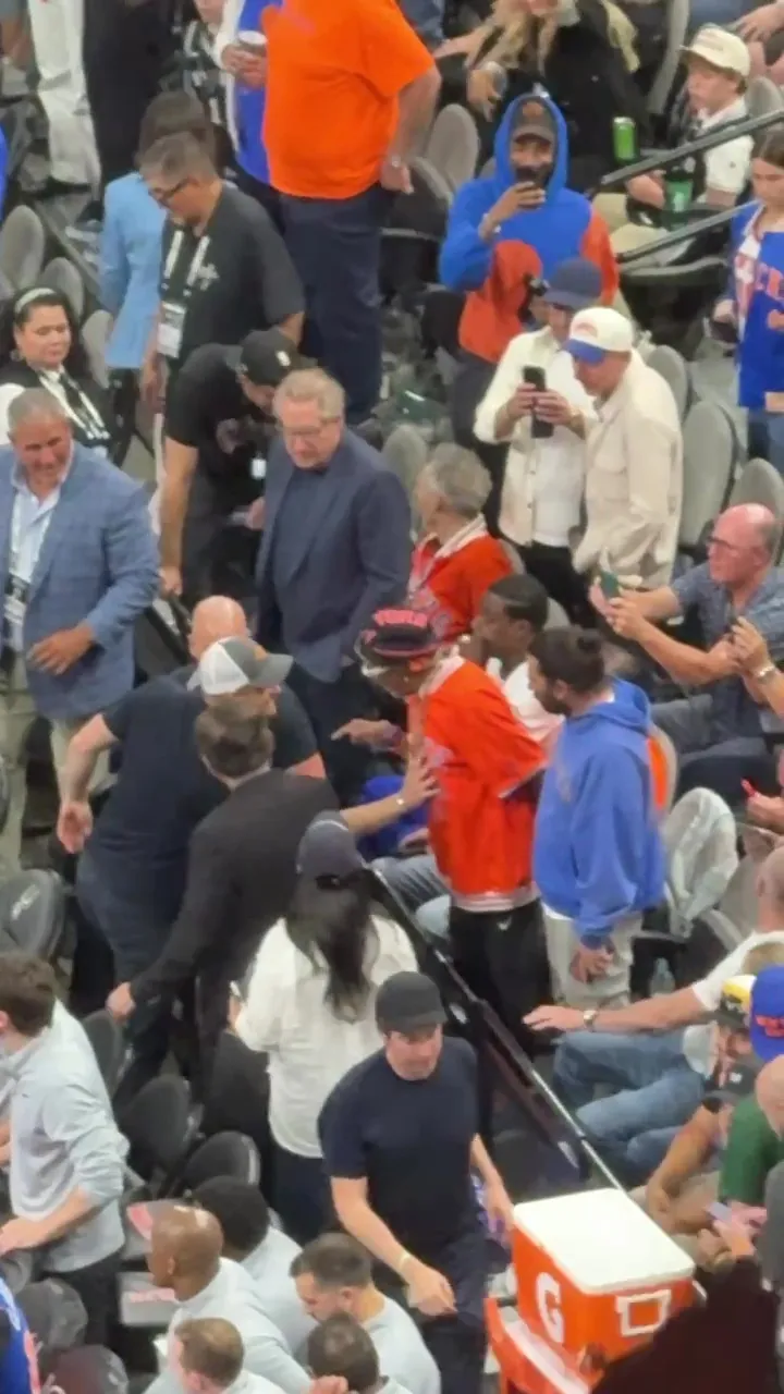 Spike Lee in an orange shirt and backwards hat denying Prince Harry's handshake in a crowded basketball arena.