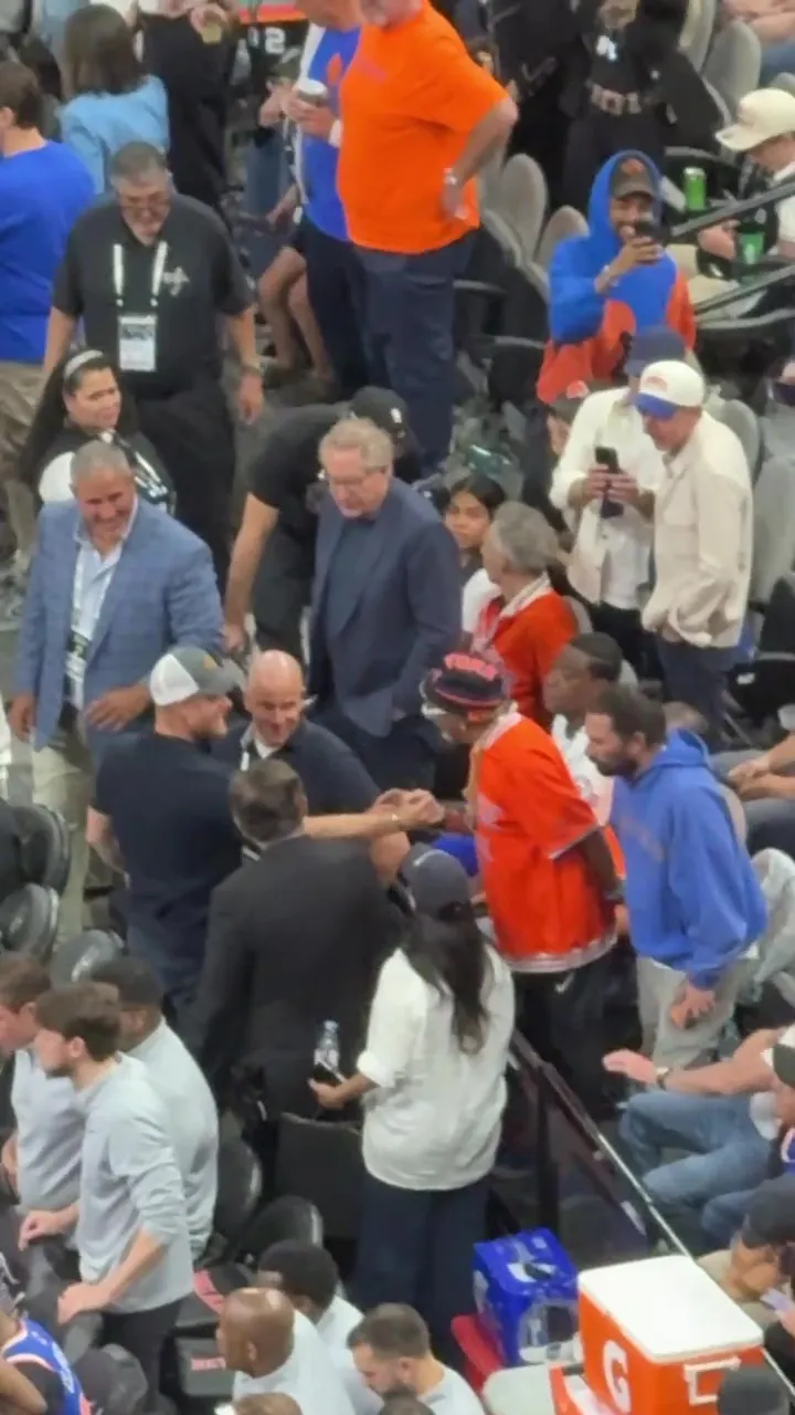 Spike Lee, in an orange t-shirt and hat, standing near Prince Harry who is dressed in a blue suit, at a basketball game.