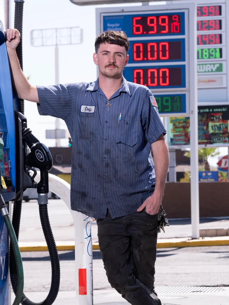 Cody, a Mobil gas station attendant in Needles, CA, stands next to a gas pump and a price sign showing gas at $6.99 per gallon.