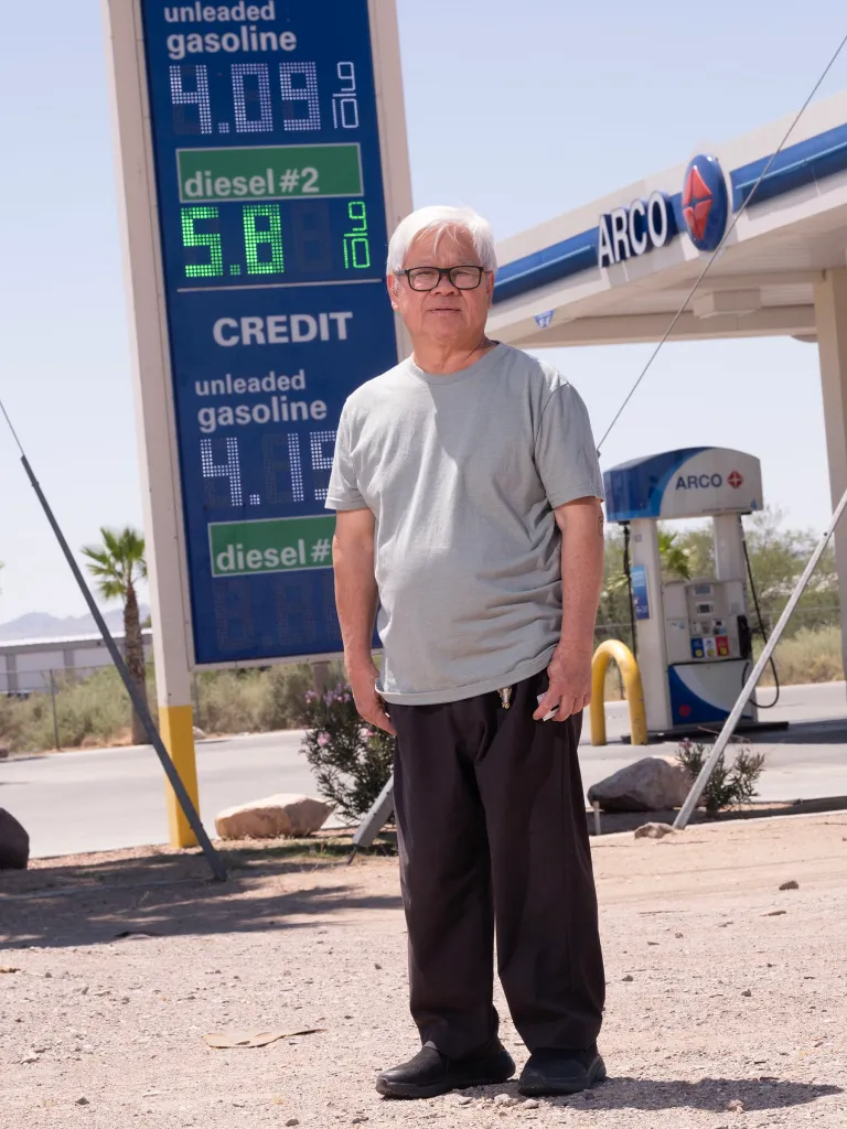 A man in glasses and a t-shirt stands in front of a gas station sign displaying unleaded gas at $4.09 9/10 and diesel #2 at $5.89 9/10.