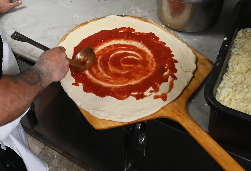 A man's hand spreads sauce on a pizza dough.