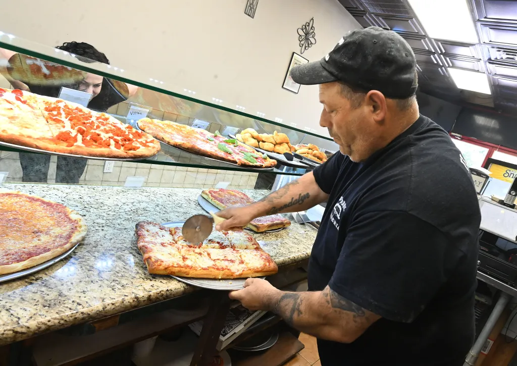 A man works behind a pizza counter and cuts into a pizza