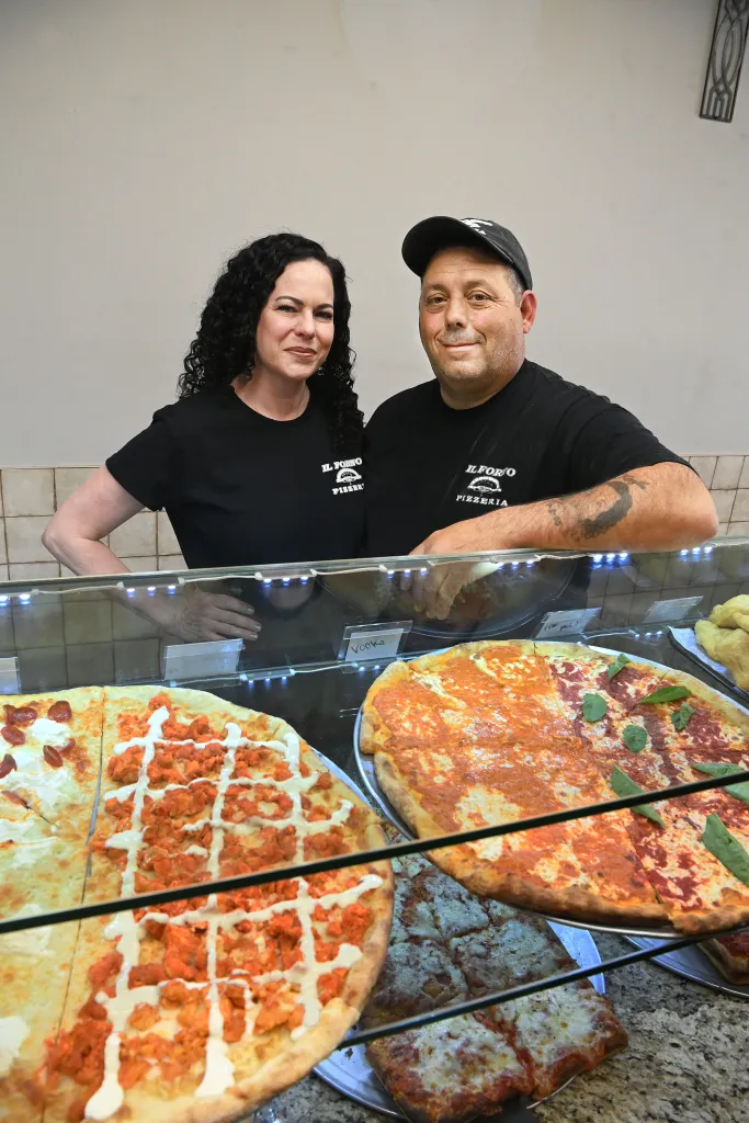 A man in his 50s standing next to a woman, his wife, both wearing black in front of a pizza counter with two pies.