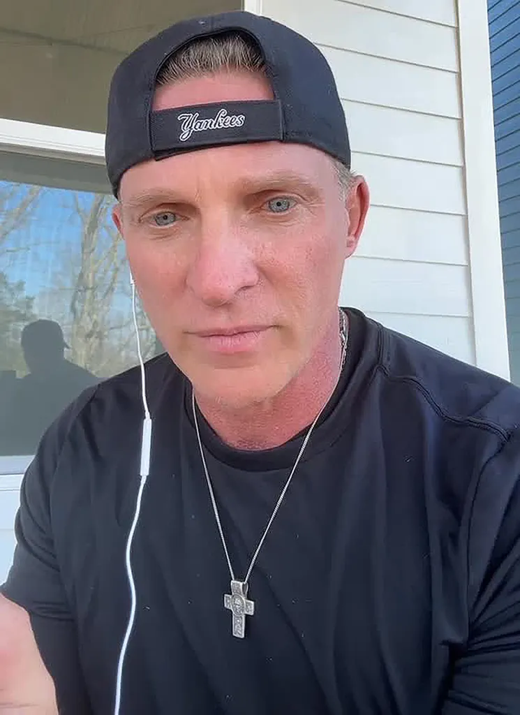 Steve Burton in a black Yankees cap and t-shirt, wearing a cross necklace and wired earbuds.