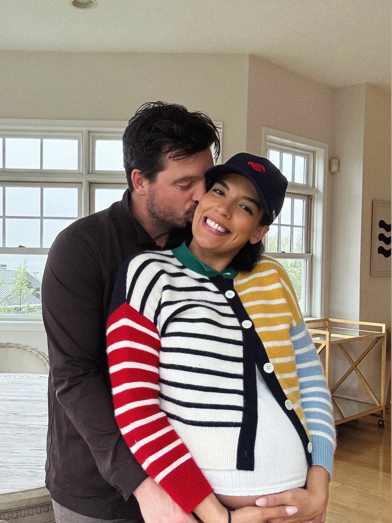 Man in a black shirt kissing the cheek of a smiling woman in a striped cardigan and baseball cap, with his arms around her baby bump.