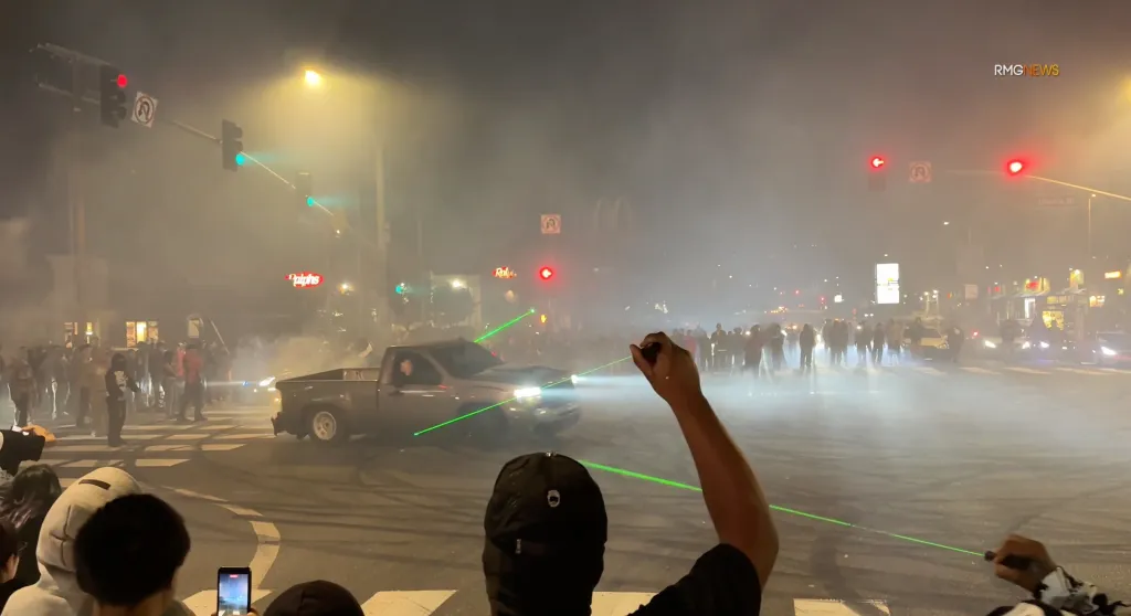 A street takeover in Baldwin Hills shows a truck doing donuts in an intersection, surrounded by a crowd and green laser beams cutting through the smoke.