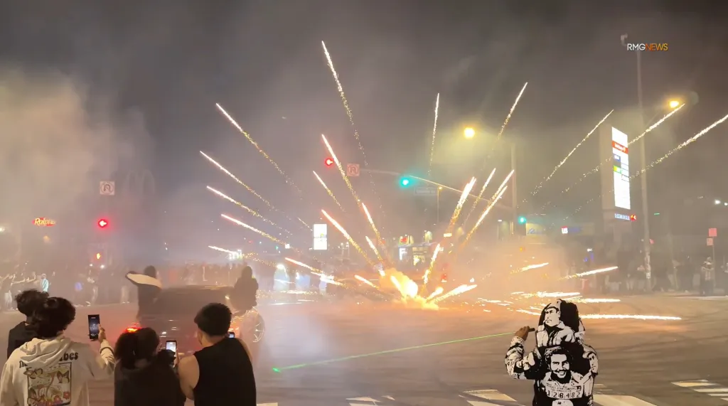 A crowd watches as fireworks explode from a car performing a street takeover at an intersection at night.