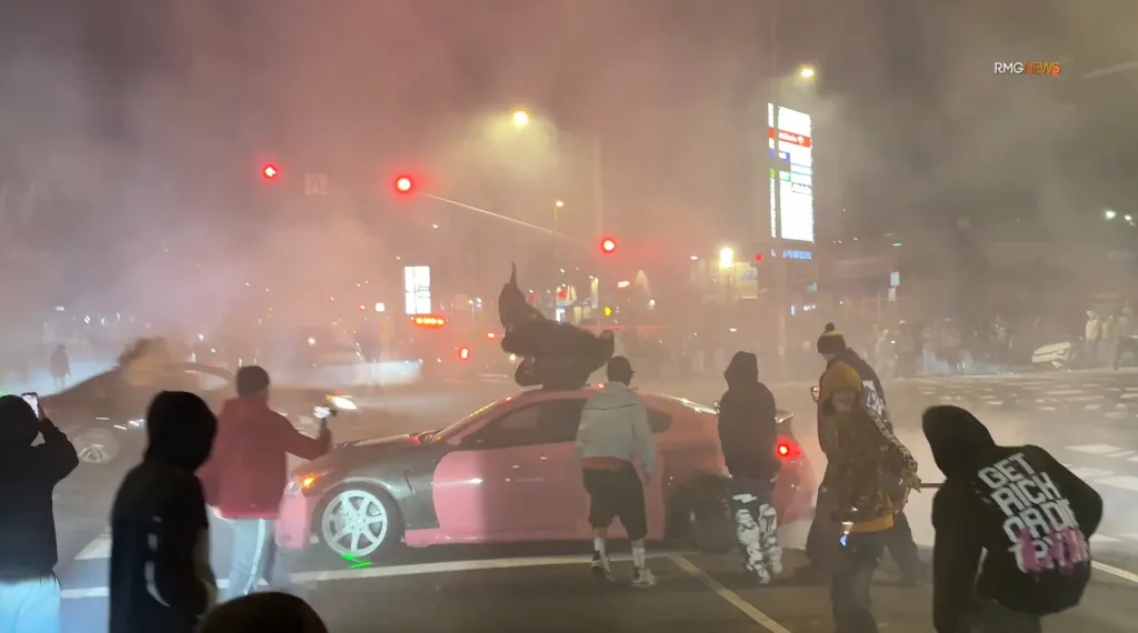 A pink and grey car at a street takeover, surrounded by people and smoke from fireworks, with red traffic lights visible.