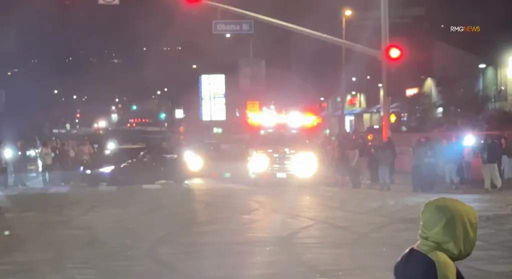 An ambulance with flashing lights is surrounded by vehicles and people at an intersection at night, with an