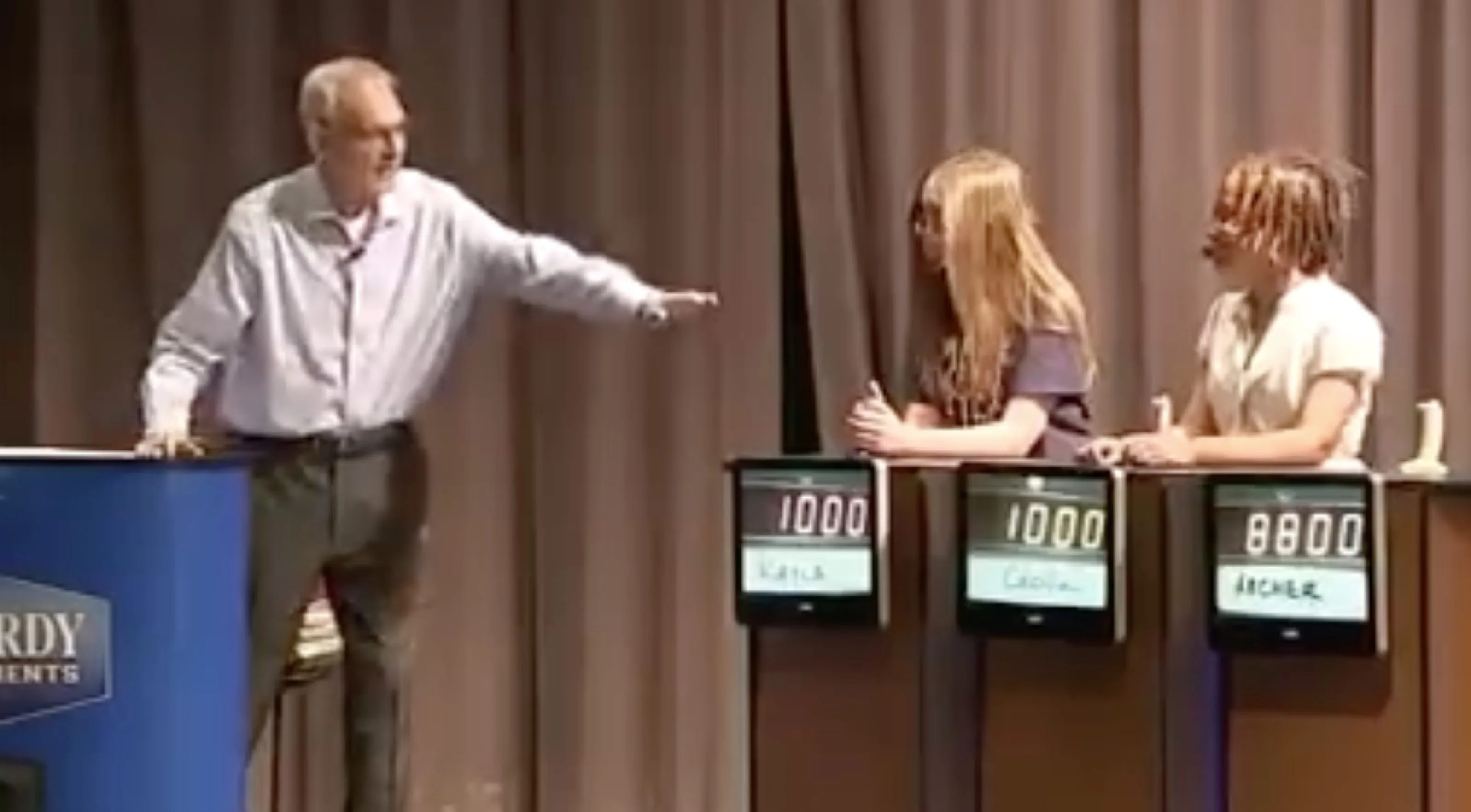 A man hosting a Jeopardy! tournament for students, with a blue podium and three student contestants behind their score displays.