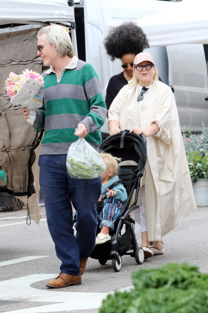 Sia, wearing a pink hat and glasses, pushes her son Somersault Wonder Bernad in a stroller next to a friend holding flowers and a bag of produce at a farmers market.