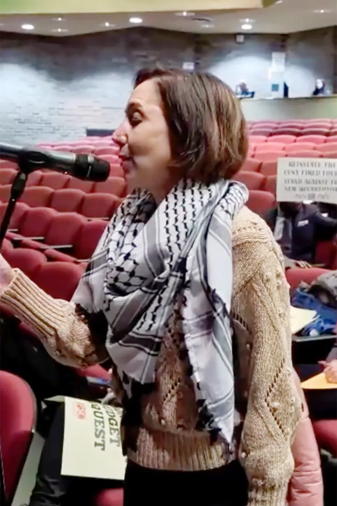 Corinna Mullin speaks into a microphone, wearing a patterned scarf and a tan sweater, with red theater seats visible behind her.