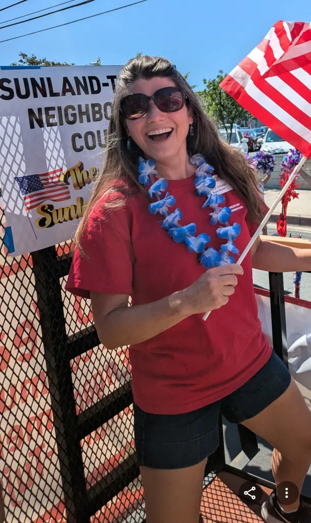 Woman wearing sunglasses and a blue lei, holding an American flag at the Sunland-Tujunga July 4th parade.