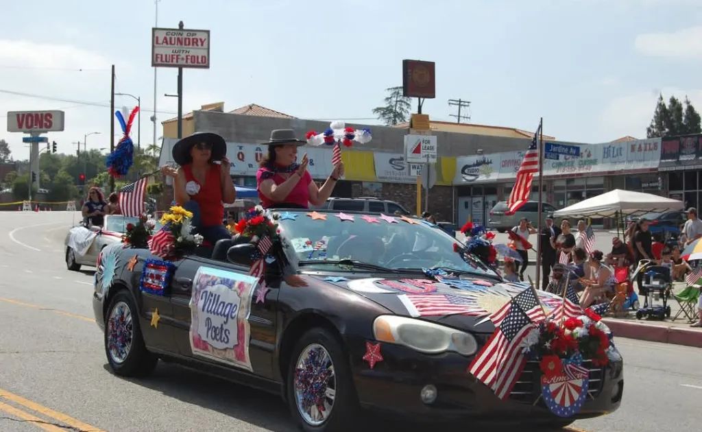A black convertible, decorated with American flags, stars, and flowers, carries two women in hats waving to parade-goers.