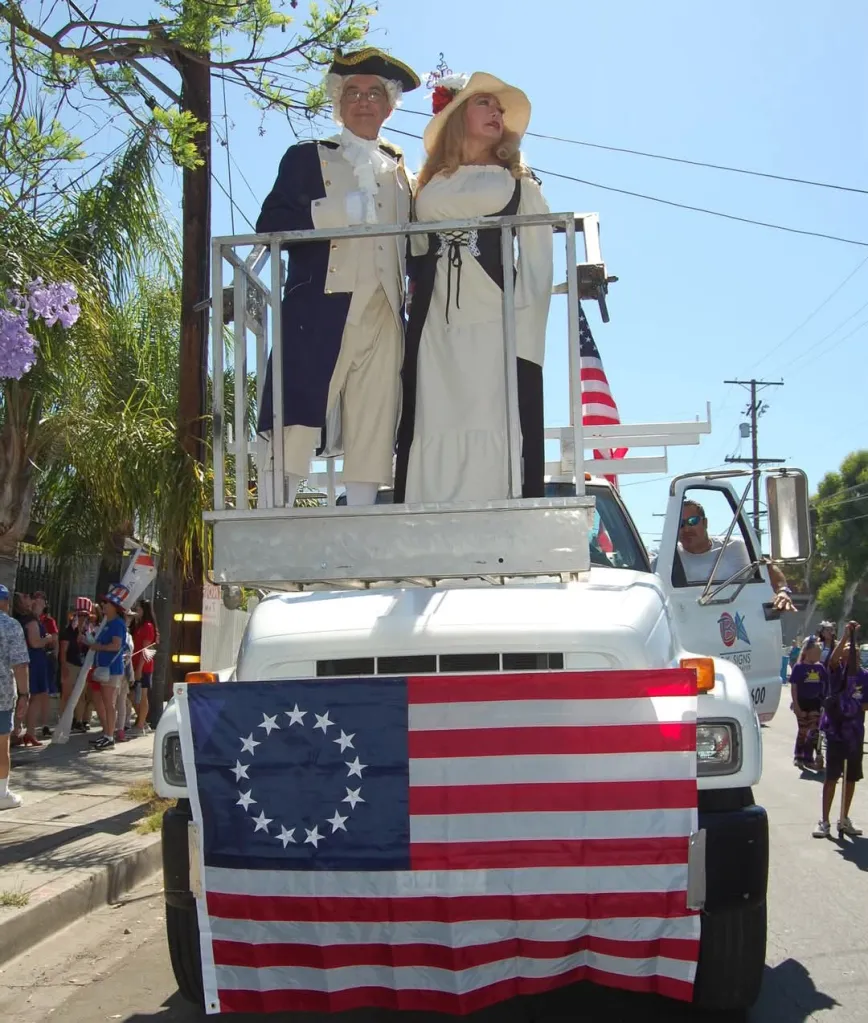 Two people in colonial attire stand on a platform mounted on a white truck with a Betsy Ross flag draped over the front, during the Sunland-Tujunga July 4th parade.