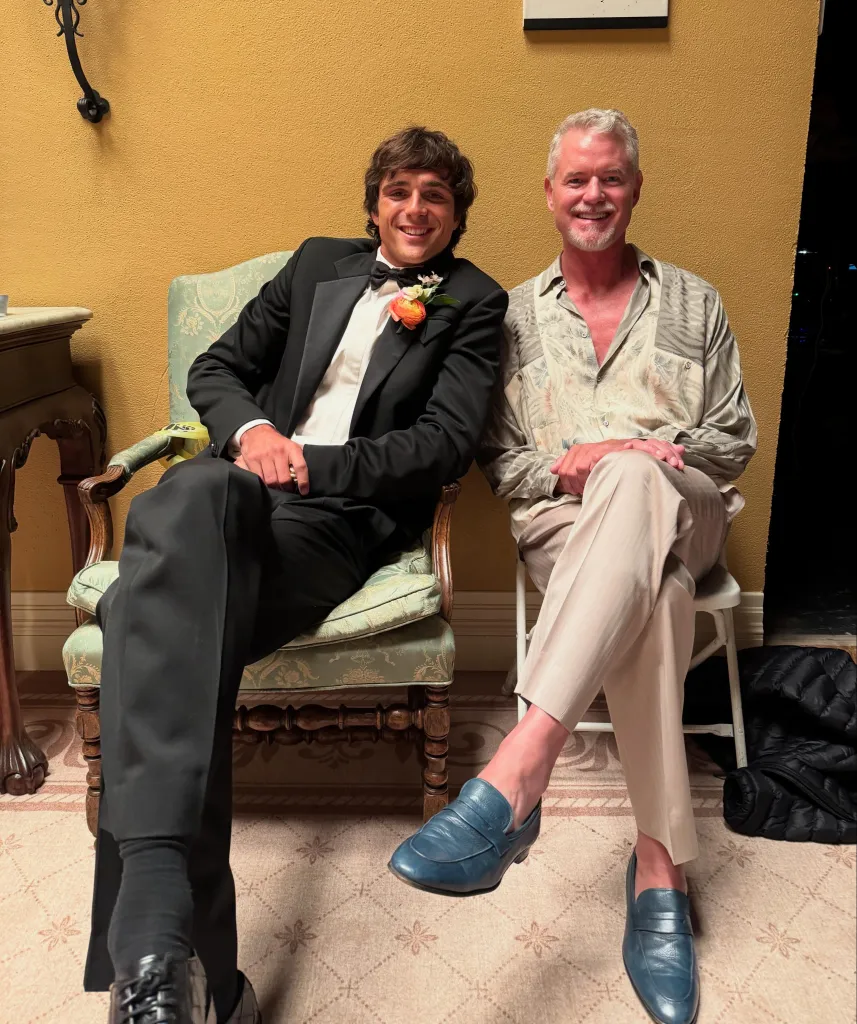 Two men sitting in chairs, one in a tuxedo and the other in a patterned shirt and beige pants.
