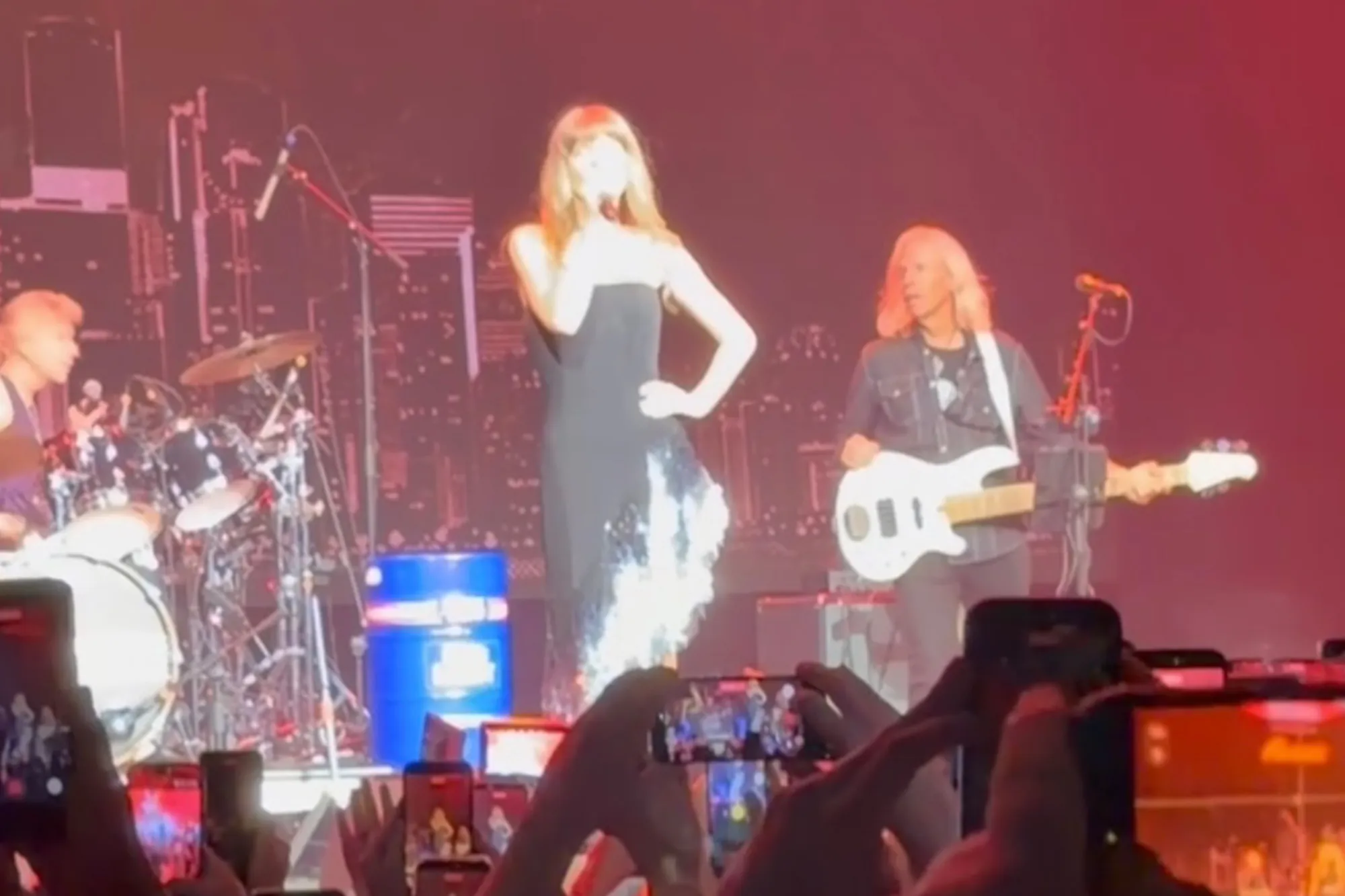 Taylor Swift performs on stage with musicians while the audience films on their phones.