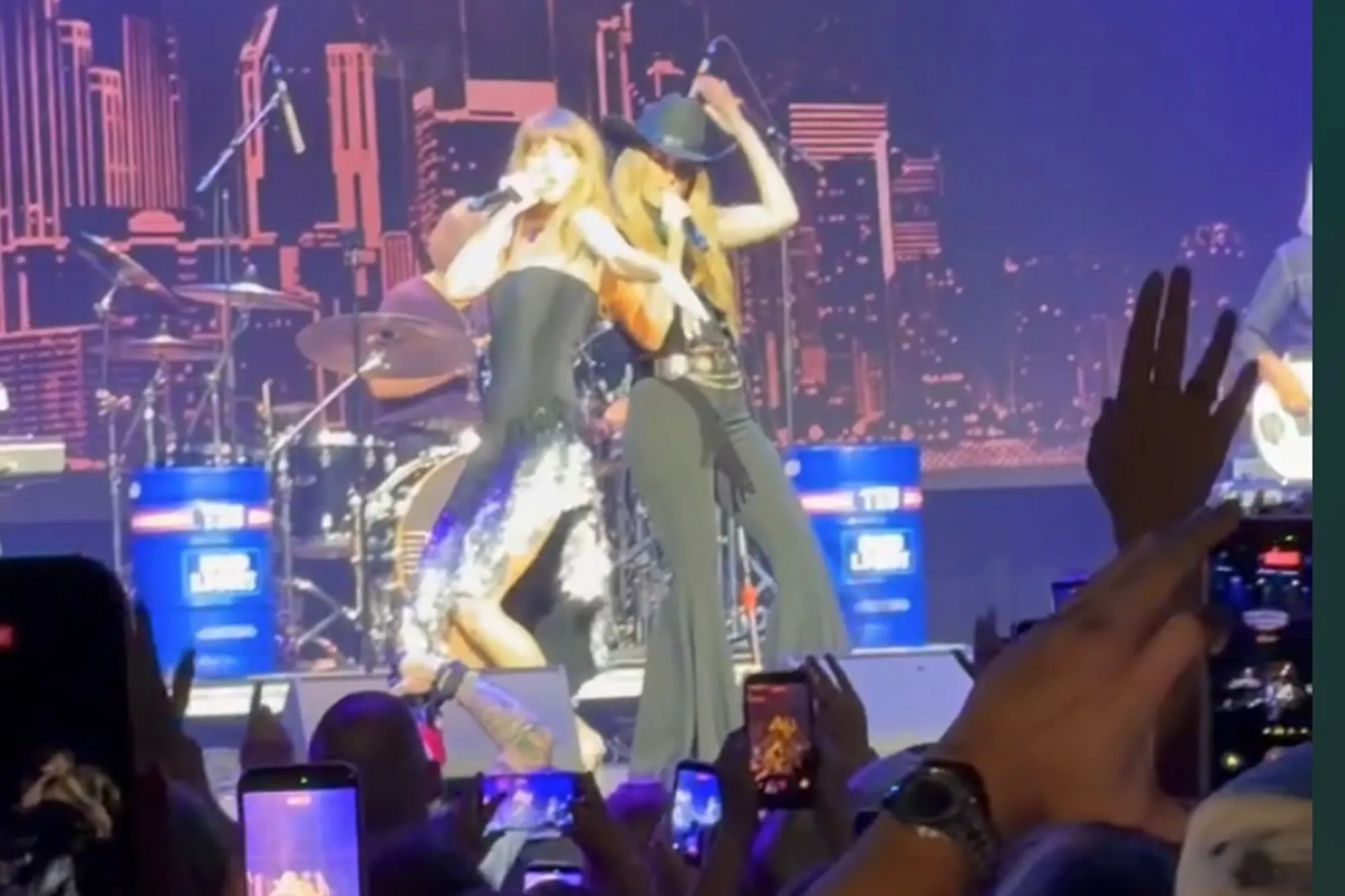 Two female singers perform on stage, with a skyline image in the background.