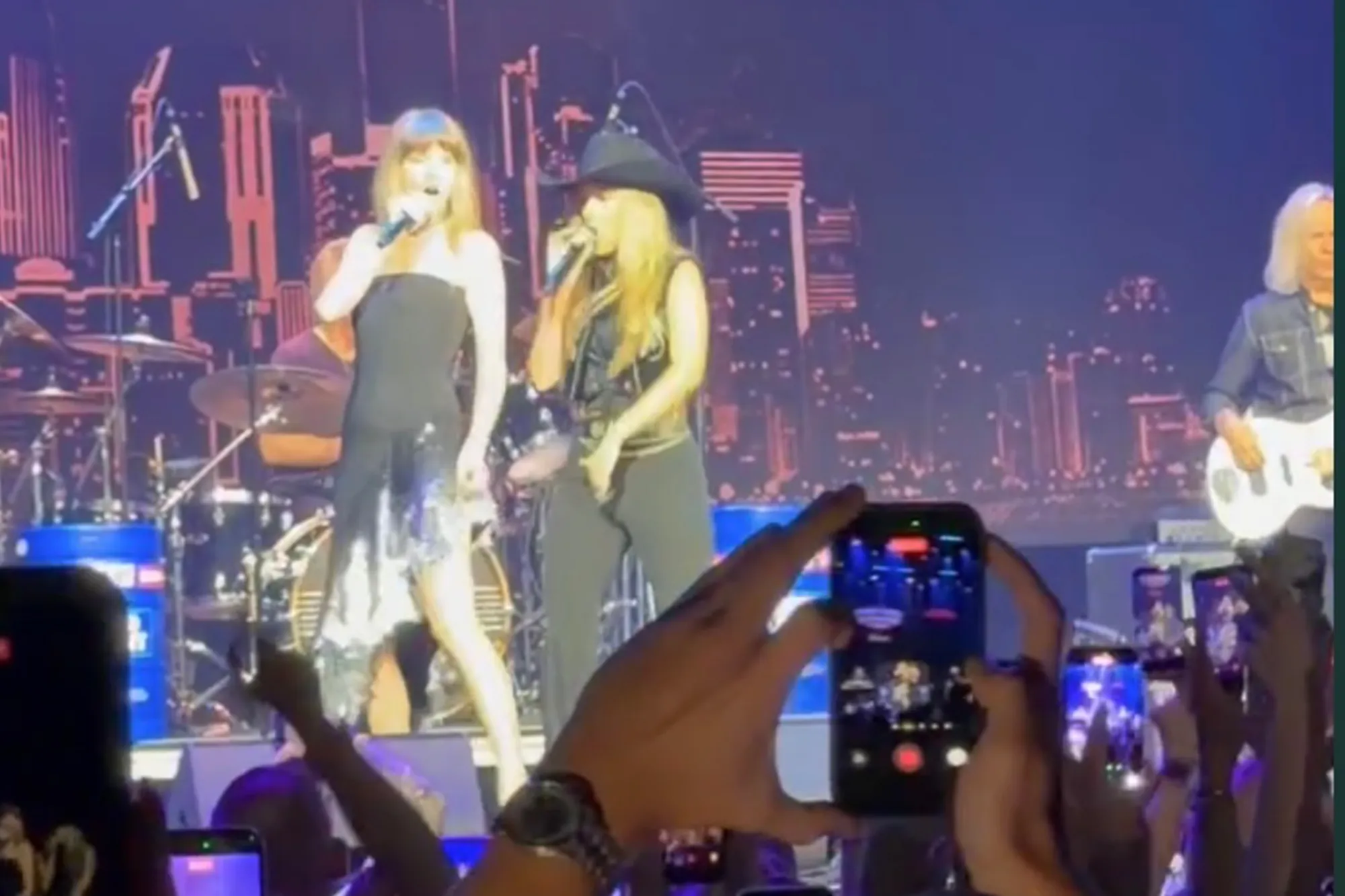 Taylor Swift singing into a microphone on stage, with a woman in a cowboy hat next to her, as concertgoers record with their phones.