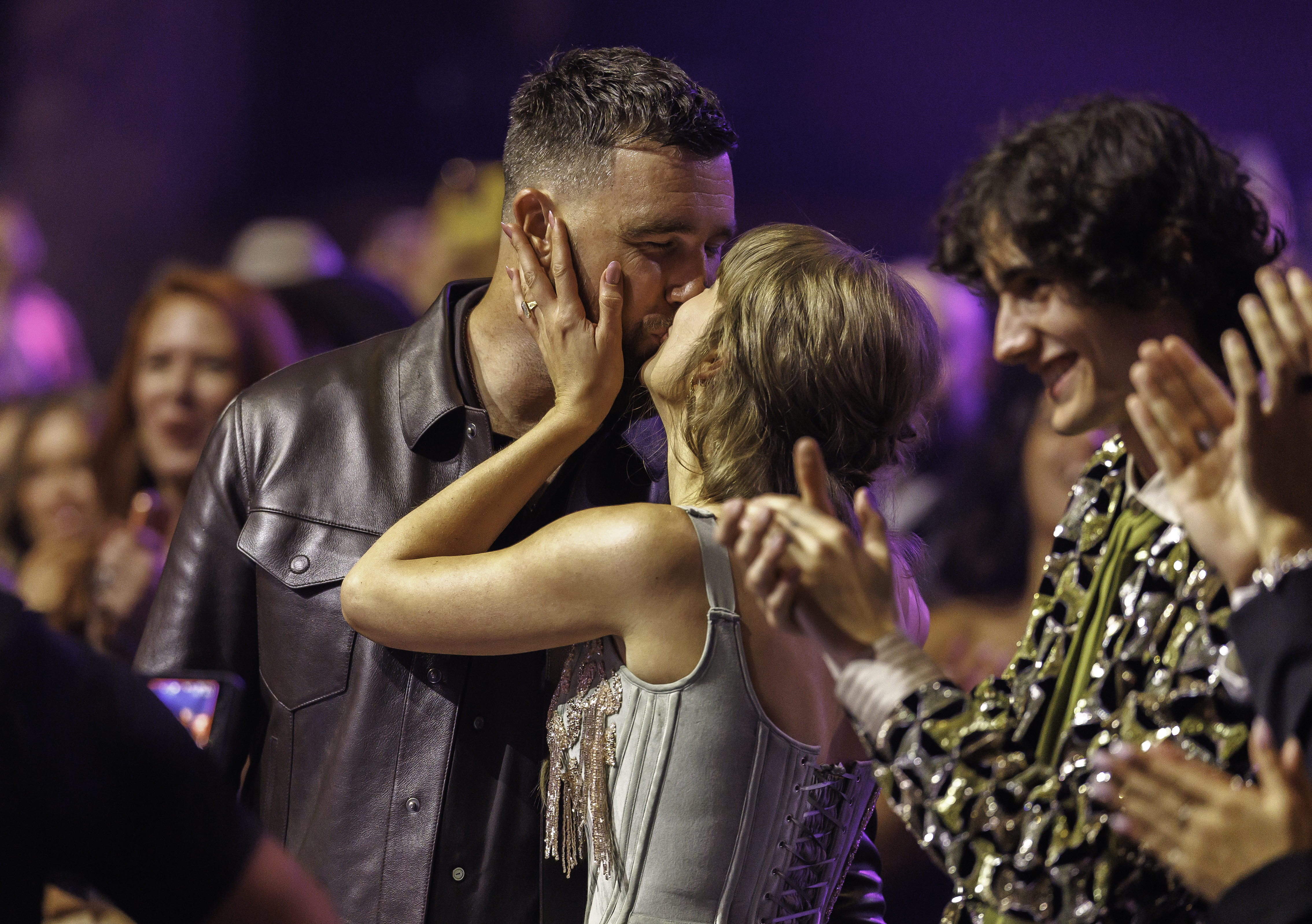 Taylor Swift kissing Travis Kelce as a man in a sequined jacket claps in the foreground.
