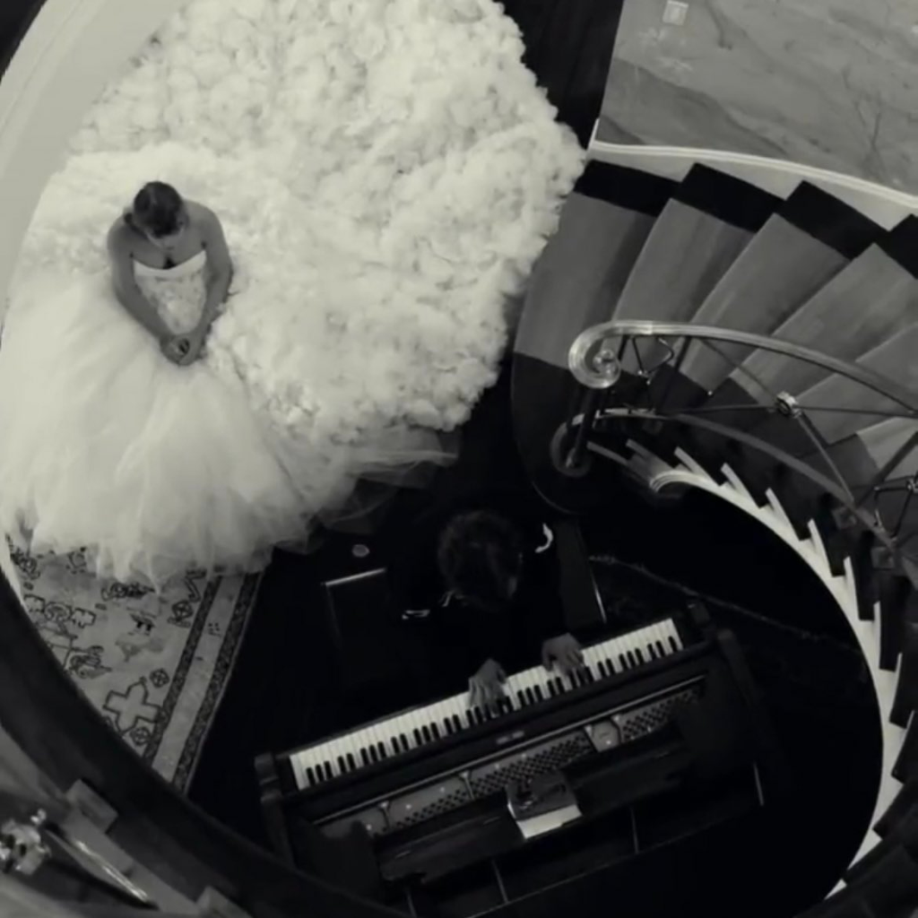 Taylor Swift in a voluminous wedding dress sitting by a piano on a spiral staircase landing.