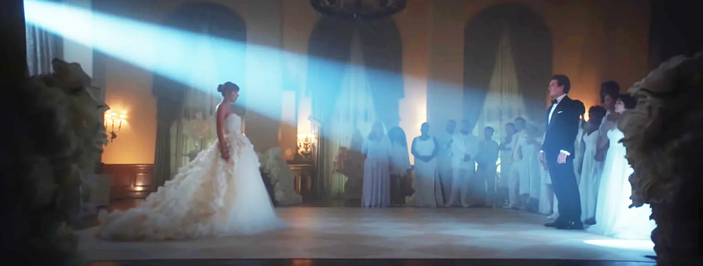 Taylor Swift in a wedding dress in a ballroom, with a man in a tuxedo and other guests in white attire.