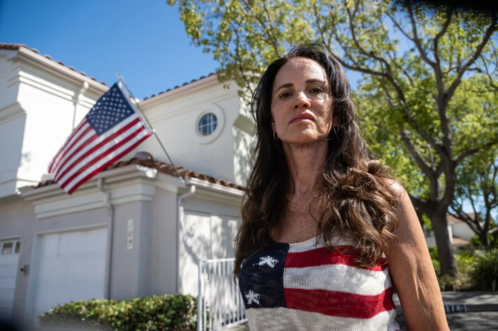 Terri Collins, wearing an American flag shirt, stands in front of her home, which displays an American flag.