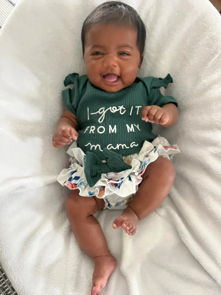 Six-month-old baby Shea smiling while lying on a white blanket, wearing a green bodysuit that says 