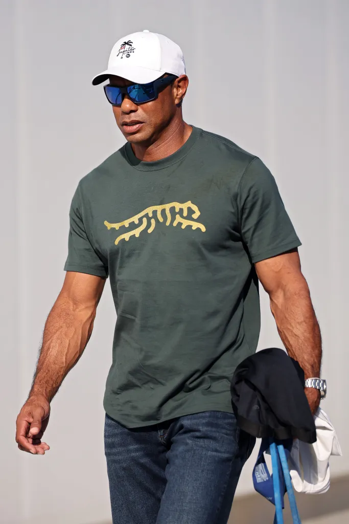 Tiger Woods wearing a dark green T-shirt with a gold tiger logo, blue jeans, sunglasses, and a white Jupiter Links Golf Club cap.