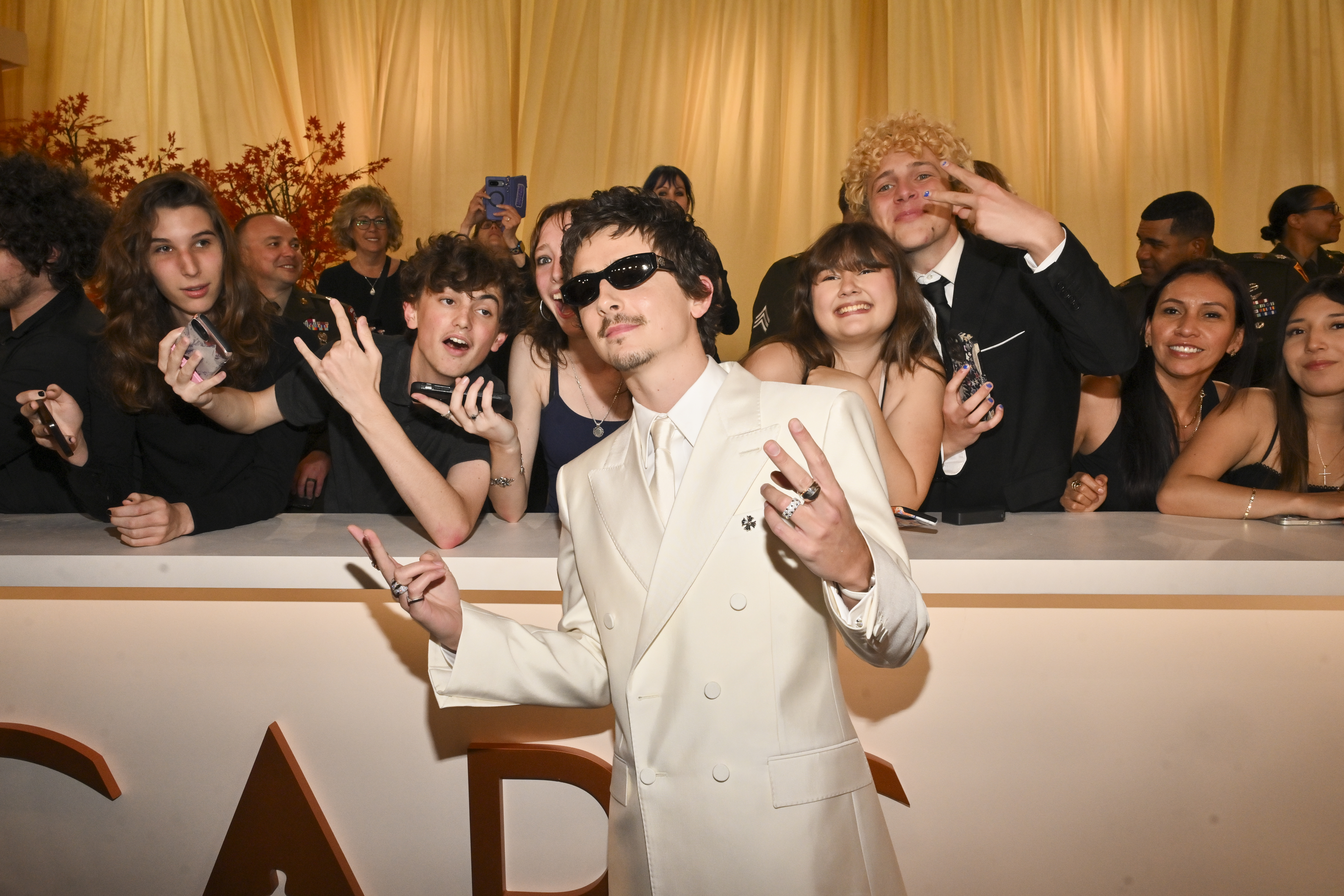 Timothée Chalamet posing in a cream suit and sunglasses with fans reaching over a barrier behind him.