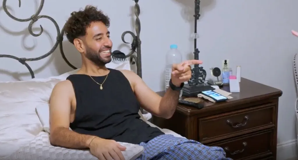 Mido sits on a bed, smiling and pointing at someone off-camera.