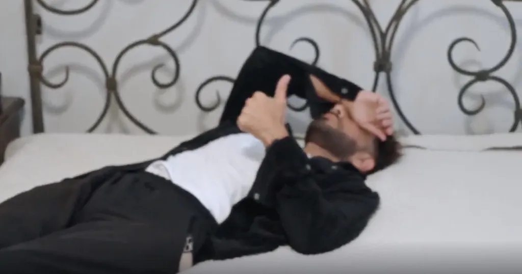 Mido lying on his back on a bed, with his right arm bent and thumb up, his left hand covering his face.