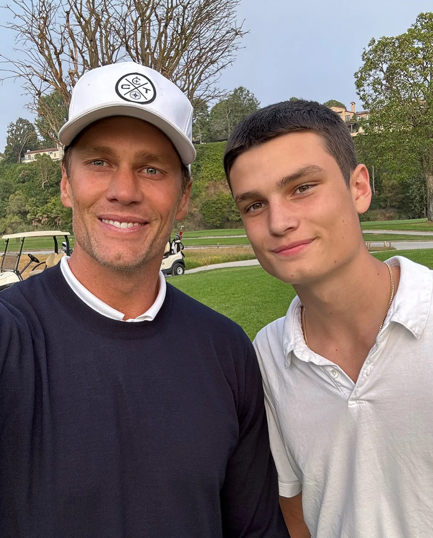 Tom Brady in a white cap and navy sweater smiles beside his son Jack, who wears a white polo shirt, on a golf course.