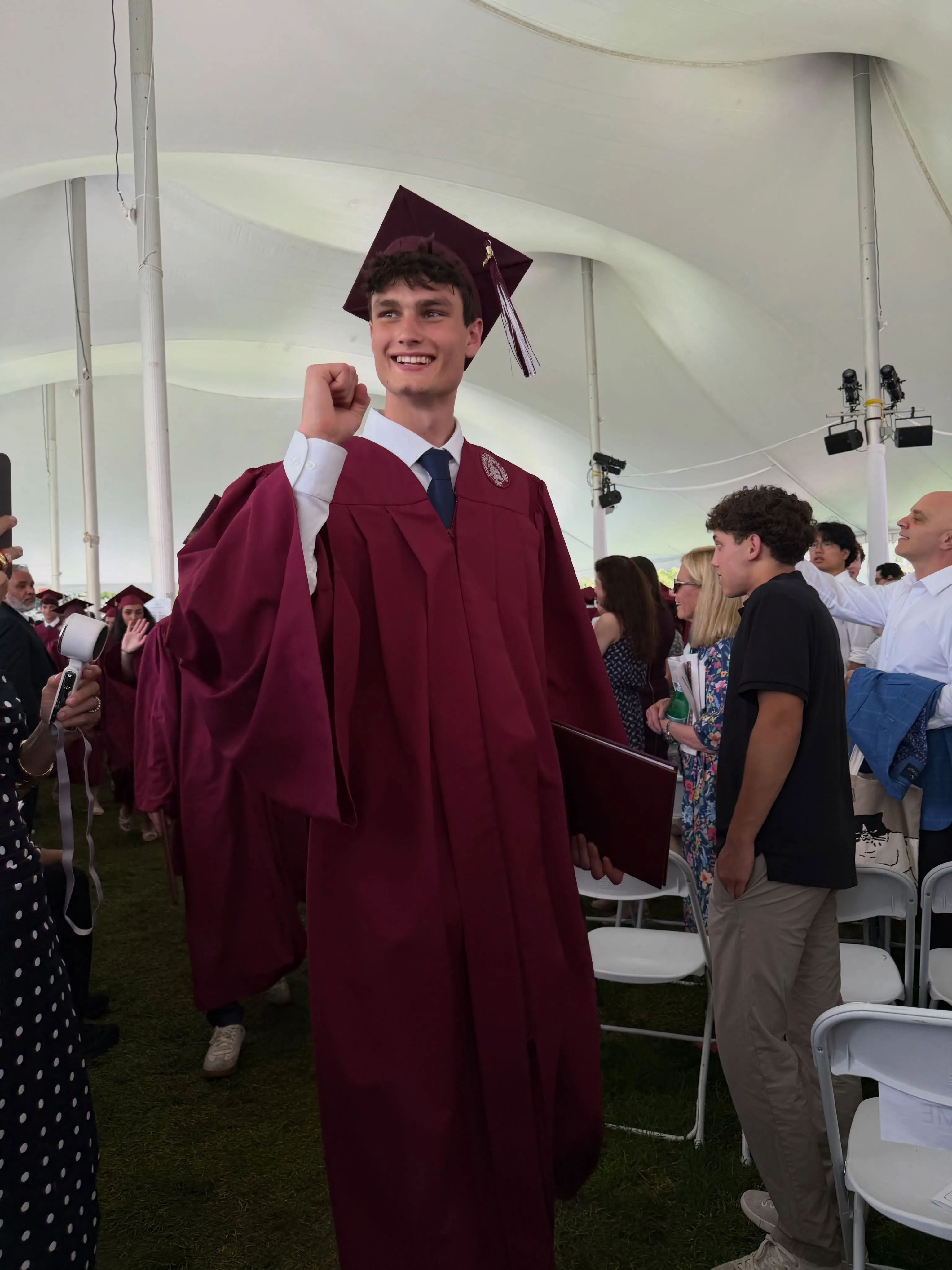 A young man in a maroon graduation gown and cap smiles and pumps his fist, holding a diploma, while walking under a white tent.