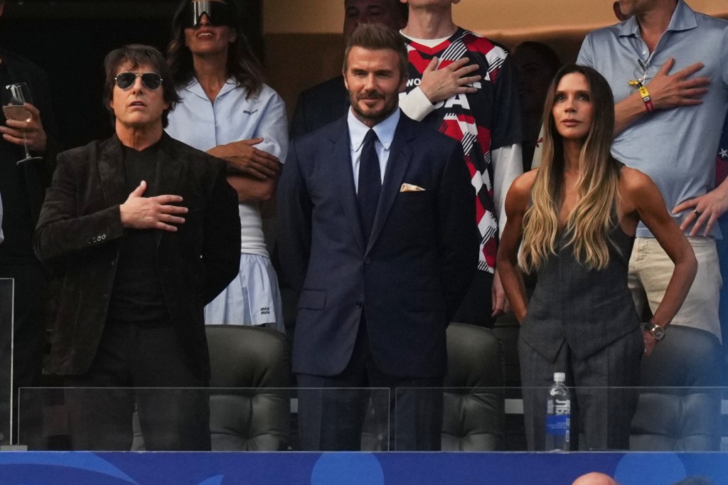Tom Cruise, in a mulberry suit, David Beckham, in a blue suit and blue tie, and Victoria Beckham, in a grey vest and grey pants, stand together in a stadium box, with their hands over their hearts.