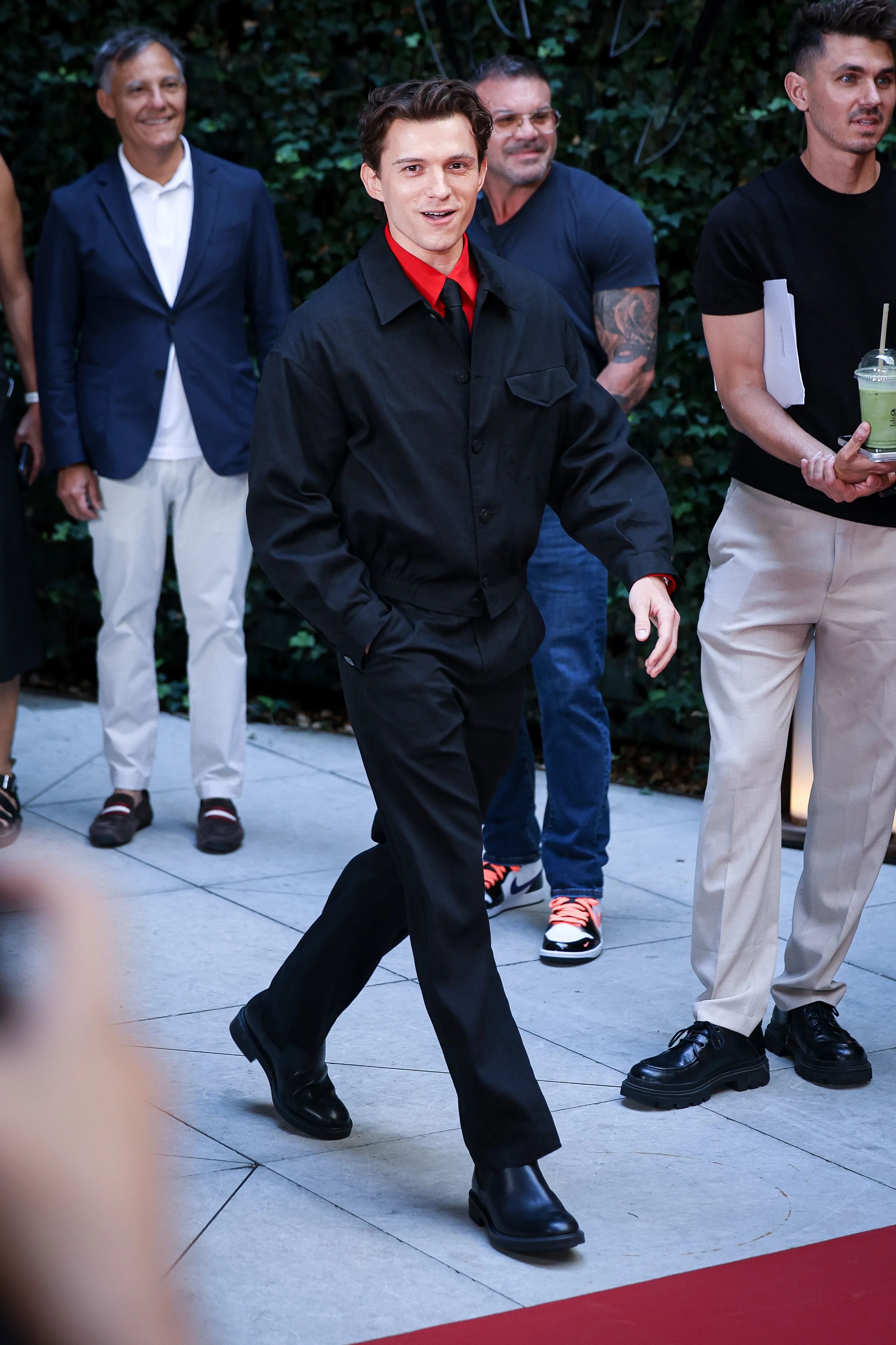 Tom Holland in a black jacket and pants over a red shirt and black tie walks down a red carpet.