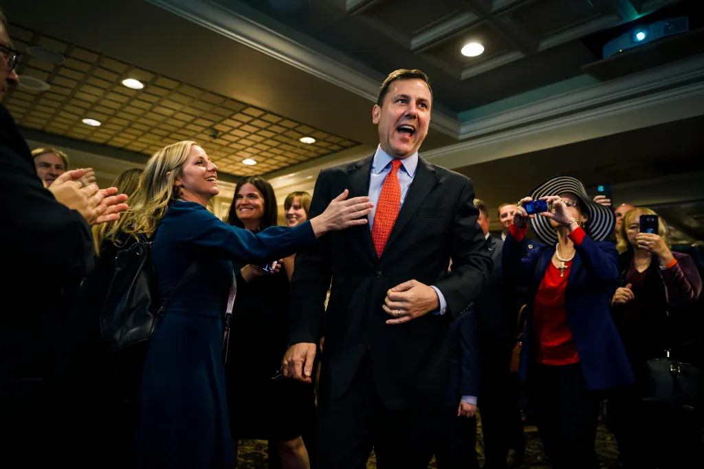 Tom Kean Jr. beams while a woman in a dark blue dress touches his arm as they are surrounded by supporters.