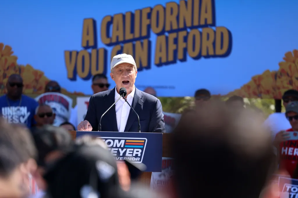 Tom Steyer speaks at a rally for Governor, with a large blue banner behind him that reads 