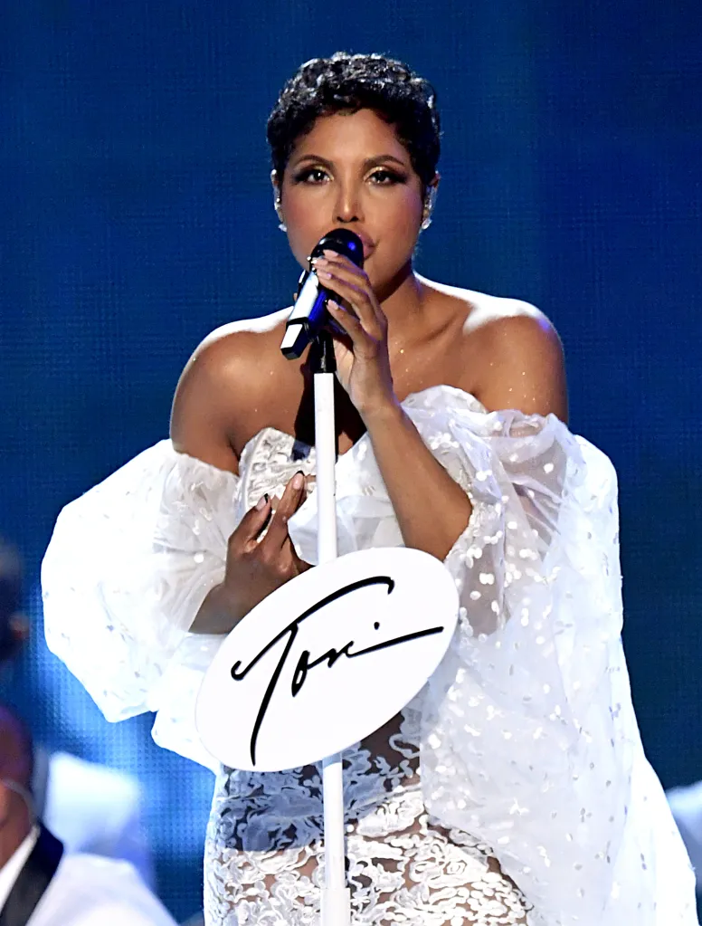 Toni Braxton performs onstage, holding a microphone with her name on a stand, wearing a white off-the-shoulder dress.