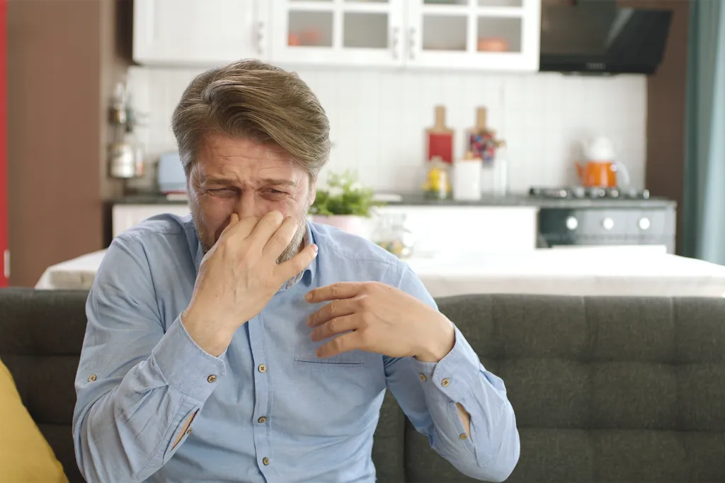 A man on a couch pinching his nose because of a bad smell.