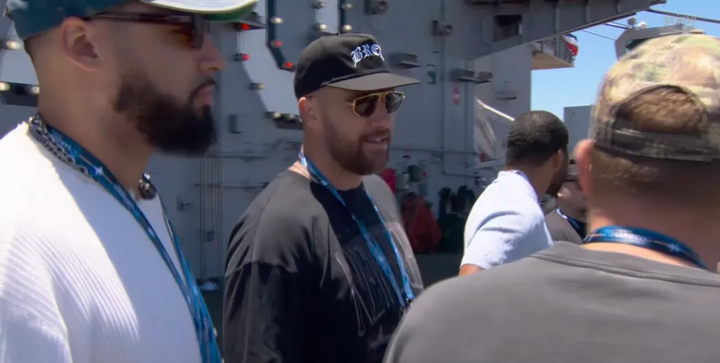 Travis Kelce with sunglasses and a baseball cap at a NASCAR event.