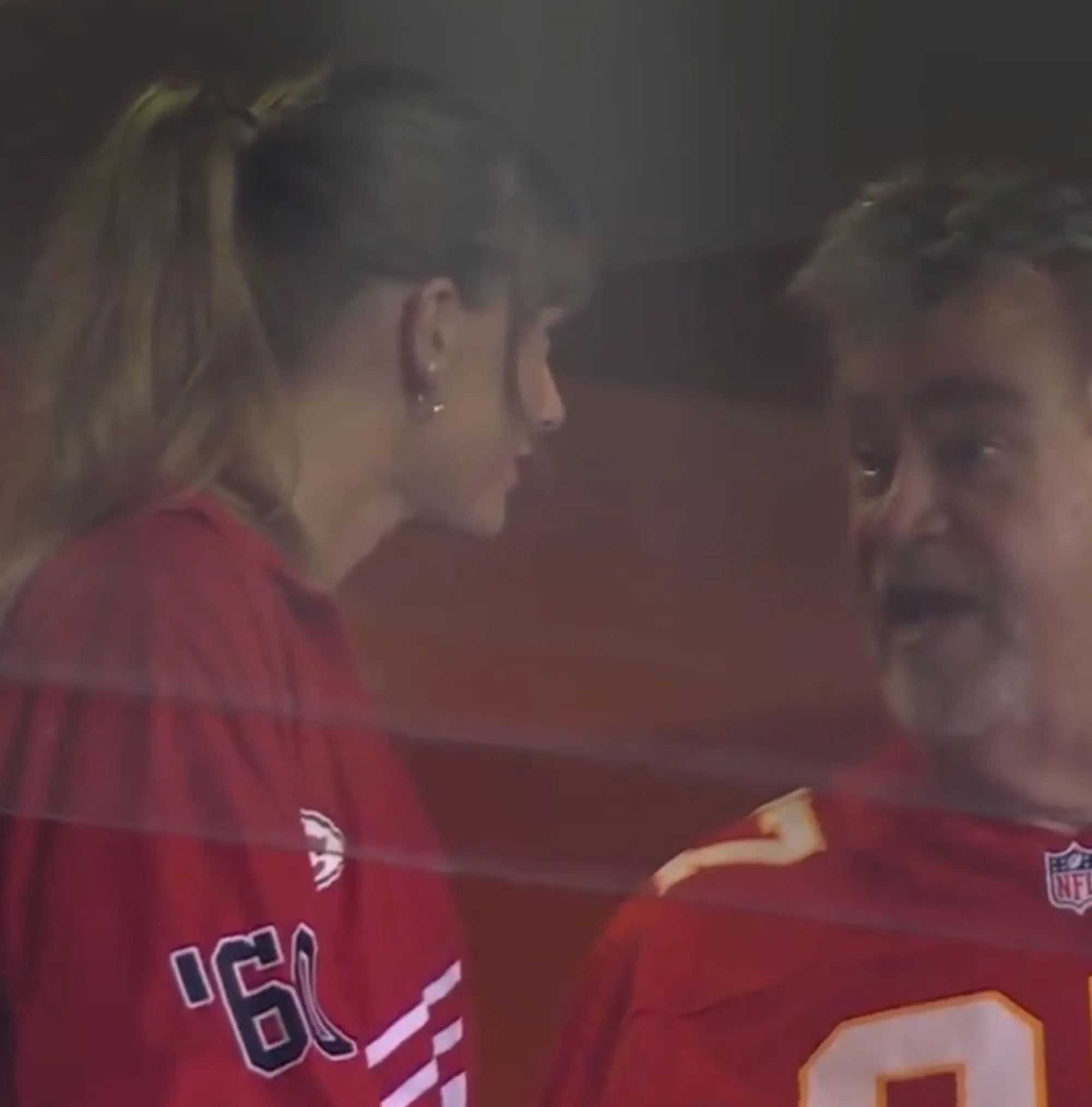 Taylor Swift in a red shirt with her hair tied back, looking towards an older man in a red football jersey.